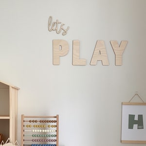May include: A wooden playroom with a white wall featuring the words "let's play" in large wooden letters. There is a wooden dollhouse, a wooden abacus, and a framed print with the letter "H" on the wall.