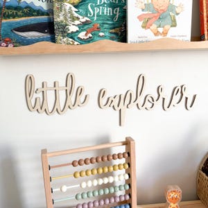 May include: Wooden wall art spelling "little explorer" in cursive script. A wooden abacus with colorful beads is in the foreground. Children's books are on a shelf above the wall art.
