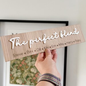 May include: A wooden sign with the words "The perfect blend" in white script. Below the text are names with heart symbols. The sign is held by a person. The background includes a framed picture.