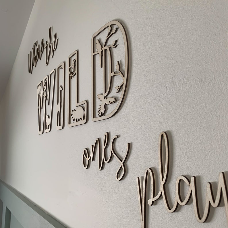 May include: Wooden wall decor with the words "Where the Wild Ones Play" in a whimsical font. The word "Wild" is decorated with cut-out images of animals and plants.