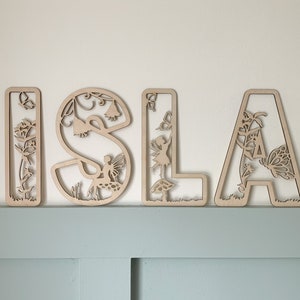 May include: Wooden letters spelling "ISLA" with a fairy and flower design. Each letter is intricately cut with a delicate floral pattern and features a fairy figure.