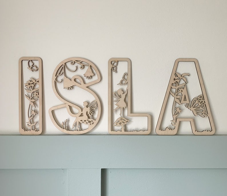 May include: Wooden letters spelling "ISLA" with a fairy and flower design. Each letter is intricately cut with a delicate floral pattern and features a fairy figure.