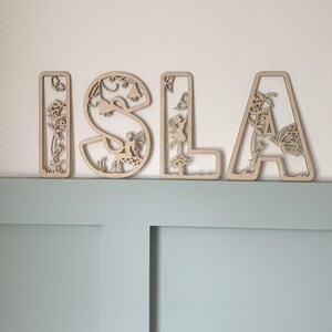 May include: Wooden letters spelling the name "ISLA" with intricate cutouts of fairies, flowers, and butterflies. The letters are a light brown color and are arranged on a light blue wall.