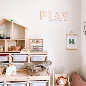 May include: A wooden playroom with a dollhouse, a wooden abacus, a wooden sign that says "Adventure Awaits", and a framed print with the letter "H". The wall has the word "PLAY" spelled out in large wooden letters.