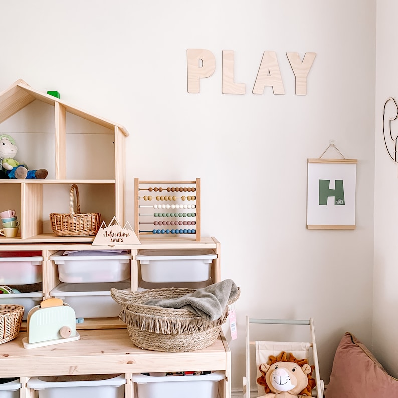 May include: A wooden playroom with a dollhouse, a wooden abacus, a wooden sign that says "Adventure Awaits", and a framed print with the letter "H". The wall has the word "PLAY" spelled out in large wooden letters.