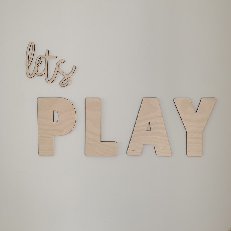 May include: Wooden wall decor with the words "let's play" in a cursive font. The letters are made of natural wood and are attached to a white wall.