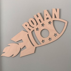 May include: A wooden rocket ship with the name "ROHAN" cut out in the front. The rocket has a flame coming out of the back and stars cut out around the body.