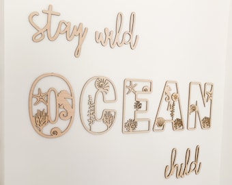 May include: Wooden wall decor with the words "stay wild ocean child" in cursive script. The word "ocean" is made up of individual letters with cut-out designs of seahorses, starfish, and coral.