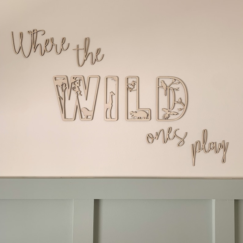 May include: Wooden wall decor with the words "Where the Wild Ones Play" cut out in a whimsical font. The letters are decorated with cut-out images of animals and plants.