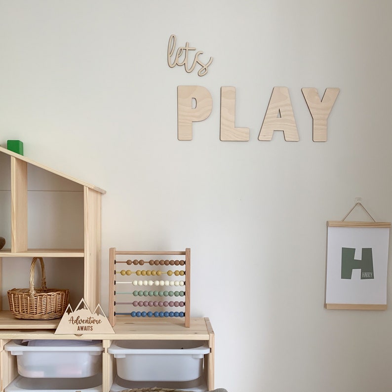 May include: A wooden playroom with a white wall featuring the words "let's play" in large wooden letters. There is a wooden dollhouse, a wooden abacus, and a framed print with the letter "H" on the wall.