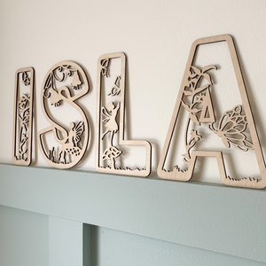 May include: Wooden letters spelling "ISLA" with a fairy and butterfly design. Each letter is intricately cut with floral and nature-inspired details.