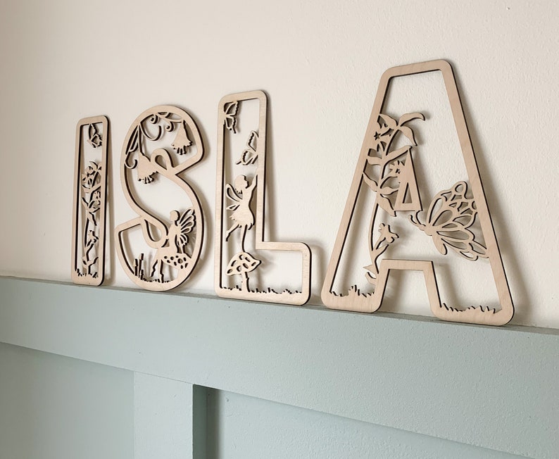 May include: Wooden letters spelling "ISLA" with a fairy and butterfly design. Each letter is intricately cut with floral and nature-inspired details.