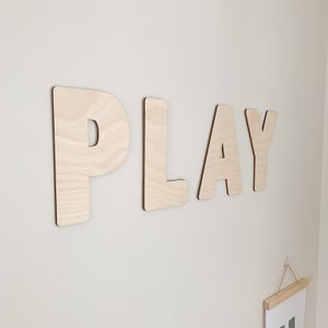 May include: Wooden letters spelling out the word "PLAY" mounted on a white wall. The letters are made of light brown wood and have a natural finish.