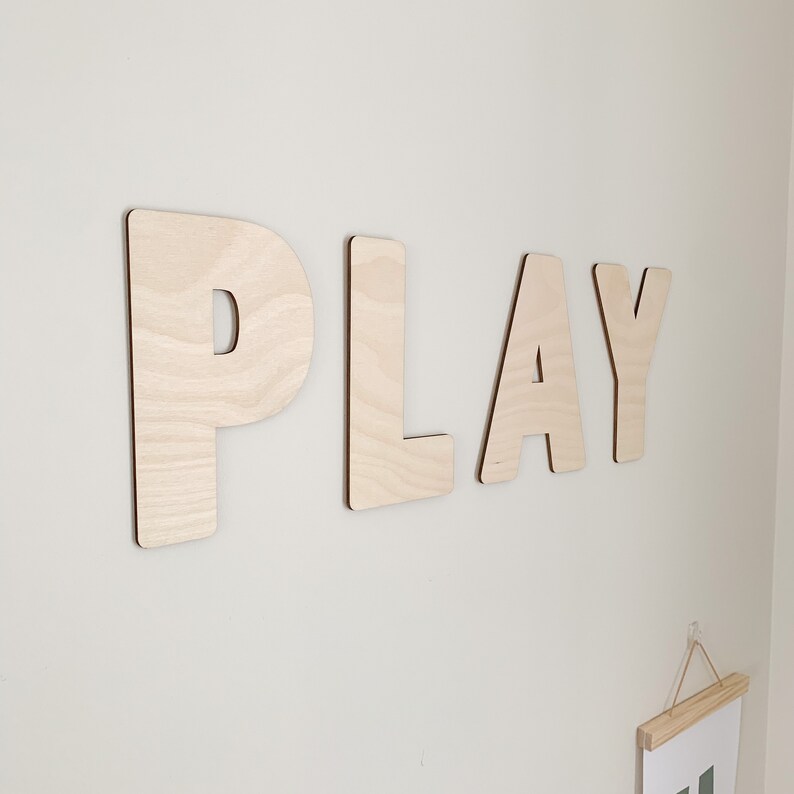May include: Wooden letters spelling out the word "PLAY" mounted on a white wall. The letters are made of light brown wood and have a natural finish.