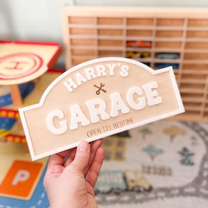 May include: A wooden sign with the text "HARRY'S GARAGE" in white, with a small wrench graphic. The sign also includes the phrase "OPEN TILL BEDTIME" in a smaller font. The sign has a light brown background and a white border.