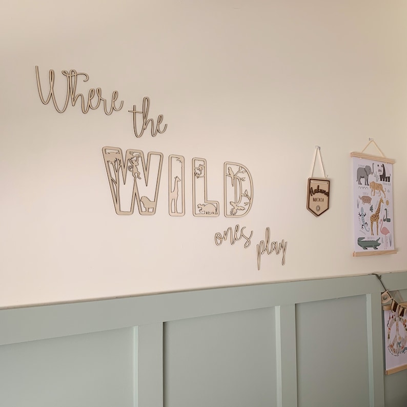 May include: A light beige wall with a wooden sign that reads "Where the Wild Ones Play". The sign is made of individual letters with cut-out animal shapes. There is a small wooden sign with the word "Remarkable" hanging on the wall. A framed print with a white background and illustrations of animals is hanging on the wall.
