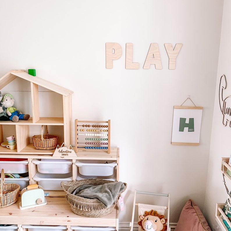 May include: A playroom with a wooden toy house, a wooden abacus, a wooden sign that says "Adventure Awaits", a wooden basket, a wooden toaster, a wooden sign that says "PLAY", a framed print with the letter "H", and a stuffed animal lion.