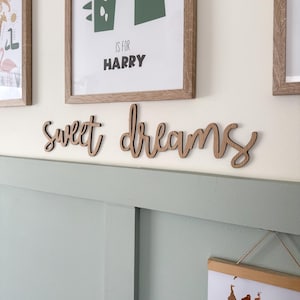 May include: A wooden sign in cursive lettering spelling out "sweet dreams". The sign is a light brown colour and is mounted on a white wall. Two framed pictures are above the sign. The wall below the sign is painted a pale green colour.