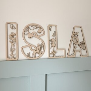 May include: Wooden letters spelling the name "ISLA" with intricate cutouts of fairies, flowers, and other nature-inspired designs. The letters are painted in a natural wood color.