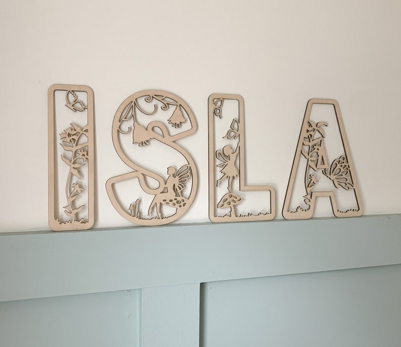 May include: Wooden letters spelling the name "ISLA" with intricate cutouts of fairies, flowers, and other nature-inspired designs. The letters are painted in a natural wood color.