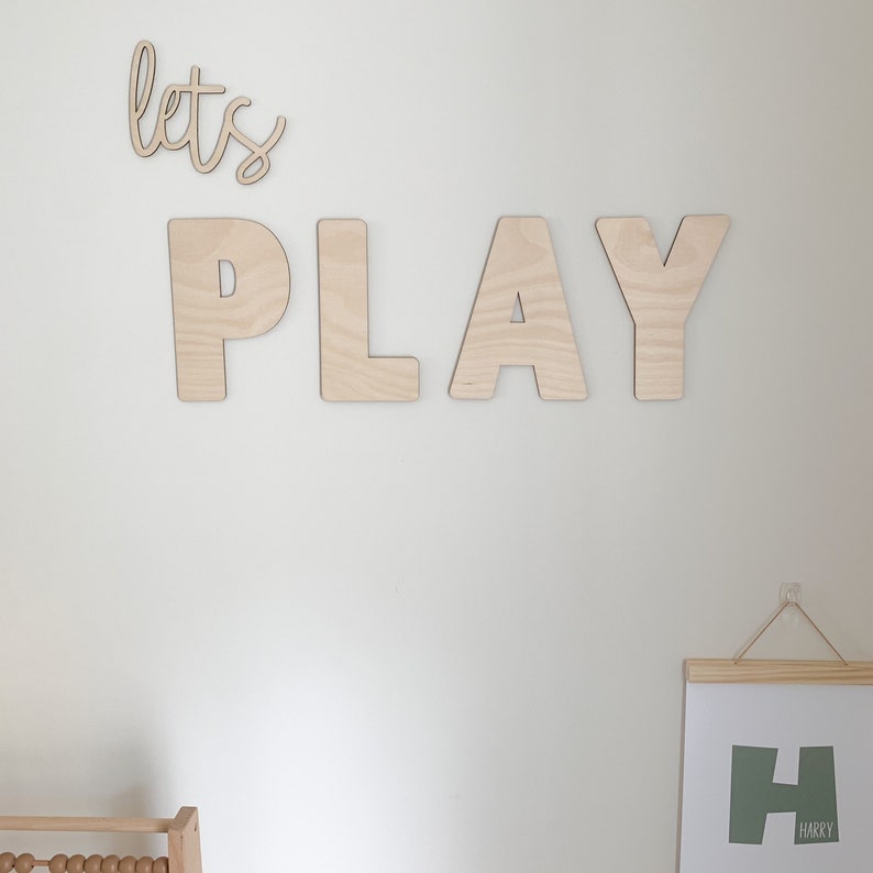 May include: Wooden wall decor with the words "let's play" in cursive script and large block letters. The letters are made of natural wood and have a light brown color. A framed print with the letter "H" and the name "Harry" is also visible in the image.