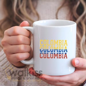 Puede incluir: Taza de cerámica blanca con la palabra "COLOMBIA" apilada tres veces en amarillo, azul y rojo, asemejándose a la bandera colombiana. La taza tiene un asa curva y está sujeta por una persona.