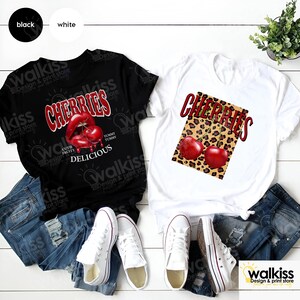 May include: Two t-shirts with a cherry design. One t-shirt is black with a red cherry lip design and the text "Cherries, Cute Fruity, Yummy Tummy, Delicious". The other t-shirt is white with a red cherry design on a leopard print background and the text "Cherries".