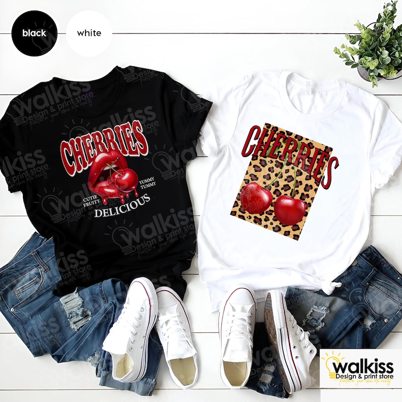 May include: Two t-shirts with a cherry design. One t-shirt is black with a red cherry lip design and the text "Cherries, Cute Fruity, Yummy Tummy, Delicious". The other t-shirt is white with a red cherry design on a leopard print background and the text "Cherries".