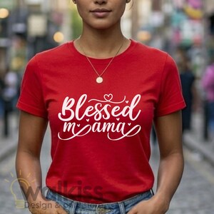 May include: A bright red short-sleeved t-shirt featuring the white text Blessed mama in a cursive script font. The design includes small heart accents integrated into the lettering. The shirt has a classic crew neckline and a standard fit.