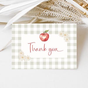 May include: A thank you card with a gingham pattern background in light green and white. The card features an apple illustration with a heart-shaped cutout and the words "Thank you" in a cursive font. Small daisy illustrations are in the corners.