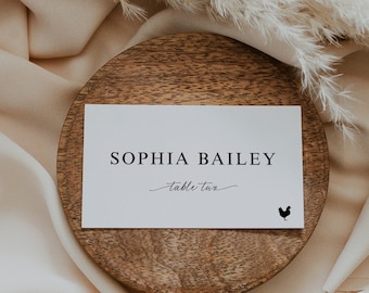 May include: White card with black text reading "Sophia Bailey, table two" with a small black rooster icon on a wooden coaster.