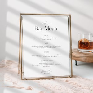 May include: A gold-framed sign with a white background and black text that reads "the Bar Menu". The menu lists beer, wine, and spirits options. The sign is sitting on a wooden table with two glasses of orange juice.