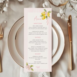 May include: A pink and white striped menu card with a floral lemon design. The menu lists appetizers, entrees, and desserts. The card is placed on a white plate with gold silverware and surrounded by flowers.