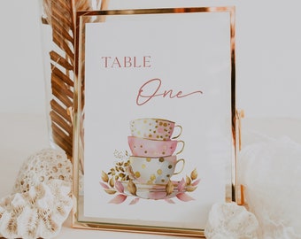 May include: Table number one with a gold frame. The number is written in cursive script. The design features a stack of three teacups with pink, gold, and white polka dot patterns. The teacups are surrounded by watercolour leaves and flowers.