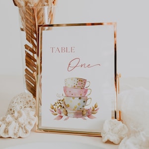 May include: Table number one with a gold frame. The number is written in cursive script. The design features a stack of three teacups with pink, gold, and white polka dot patterns. The teacups are surrounded by watercolor leaves and flowers.
