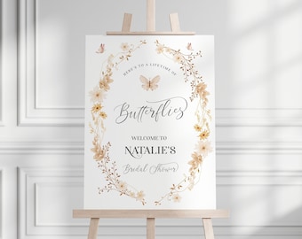 May include: A white sign with a floral wreath and a butterfly design. The text reads "Here's to a lifetime of butterflies. Welcome to Natalie's Bridal Shower."