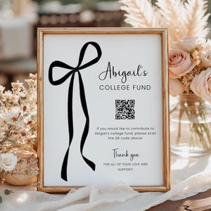May include: A framed sign with a gold border features the text "Abigail's COLLEGE FUND" and a black bow graphic. Below is a QR code and the words "Thank you" with additional text. The sign is displayed on a table with floral arrangements.