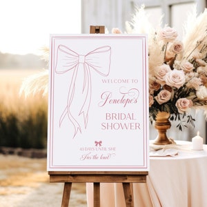May include: A white sign with a pink bow illustration and the text "Welcome to Penelope's Bridal Shower 43 days until she ties the knot".