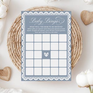 Könnte beinhalten: Eine blaue Babyparty-Bingo-Karte mit einem weißen Spitzenrand. Die Karte hat 25 Felder mit leeren Feldern, in die die Gäste ihre Vermutungen darüber eintragen können, welche Geschenke die werdende Mutter erhalten wird. Der Text "Baby Bingo" steht oben auf der Karte, und der Text "Was wird die werdende Mutter bekommen?" steht darunter.