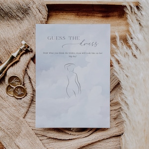 Può includere: Un biglietto bianco con uno sfondo acquerellato blu chiaro e il testo "GUESS THE dress" in corsivo nero. Sotto il testo c'è un disegno a linea di un corpo di donna con il testo "Draw what you think the bride's dress will look like on her big day!" in testo nero.