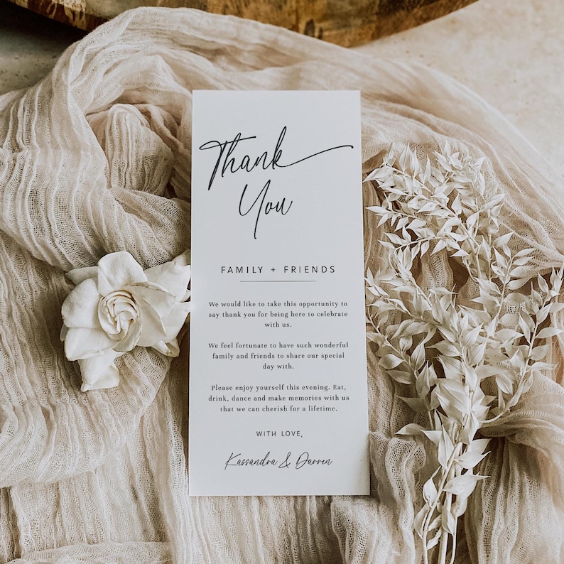K&ouml;nnte beinhalten: Eine vertikale wei&szlig;e Karte mit den Worten "Thank You" in eleganter Schrift, gefolgt von "FAMILY + FRIENDS". Die Karte enth&auml;lt eine Dankesbotschaft und ist von wei&szlig;en Blumen und einem beigen Stoffhintergrund umgeben. Es ist eine Hochzeits- oder Event-Dankeskarte.