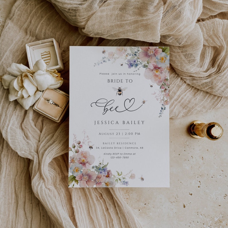 May include: A white bridal shower invitation with a watercolor floral design featuring pink, purple, and blue flowers. The text reads "Please join us in honoring Bride to Bee Jessica Bailey August 23 | 2:00 PM Bailey Residence 34 LoCosto Drive | Canmore, AB Kindly RSVP to Emma at 123-456-7890"