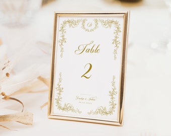 Könnte beinhalten: Goldene Tischnummerkarte mit floralem Design und dem Text "Tisch 2" und "Jenelope & Julian 08.06.20XX".