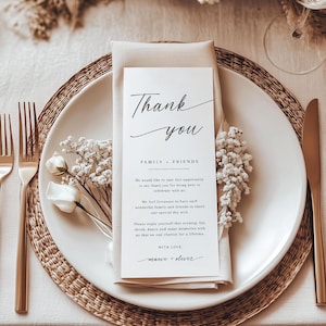 Pode incluir: Uma mesa elegante com um prato branco, talheres dourados e um guardanapo bege dobrado. Um cart&atilde;o branco com escrita preta diz "Obrigado" e inclui uma mensagem para familiares e amigos. Flores brancas e um sousplat completam o conjunto.