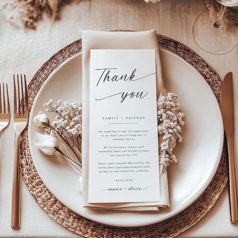 Pode incluir: Uma mesa elegante com um prato branco, talheres dourados e um guardanapo bege dobrado. Um cart&atilde;o branco com escrita preta diz "Obrigado" e inclui uma mensagem para familiares e amigos. Flores brancas e um sousplat completam o conjunto.