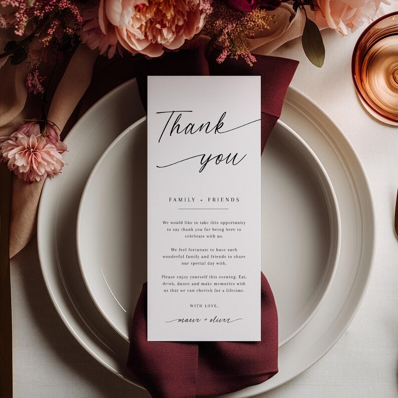 Pode incluir: Um lugar de recep&ccedil;&atilde;o de casamento branco com um guardanapo bord&ocirc; e um cart&atilde;o de "Obrigado". O cart&atilde;o tem uma escrita elegante e diz "Obrigado, Fam&iacute;lia + Amigos". Arranjos florais e vidraria ao fundo.