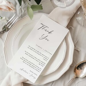 May include: A white place setting with a folded napkin, a fork, and a spoon. A white card with the words "Thank You" in elegant script is placed on the plate. The card also includes a message about celebrating with family and friends.