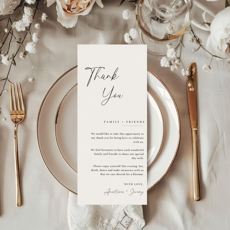 May include: A wedding table setting with a gold fork, knife, and a white plate with a gold rim. A cream-colored card with the words "Thank You" and a message is placed on the plate. White flowers and a glass of water are in the background.