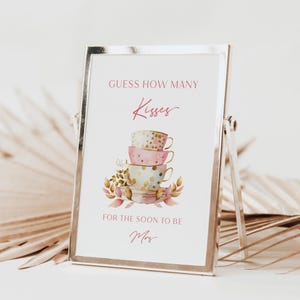 May include: A white card with pink and gold accents in a silver frame. The card says "Guess how many Kisses for the soon to be Mrs." and features a stack of four teacups with pink, gold, and white polka dots.