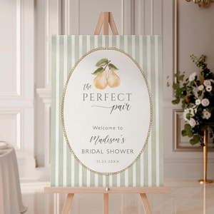 Peut inclure: Panneau de bienvenue sur chevalet en bois avec fond à rayures vertes et blanches. Le panneau comporte un cadre ovale avec les mots "the PERFECT pair" et "Welcome to Madison's BRIDAL SHOWER" en écriture élégante, avec une illustration de deux poires.